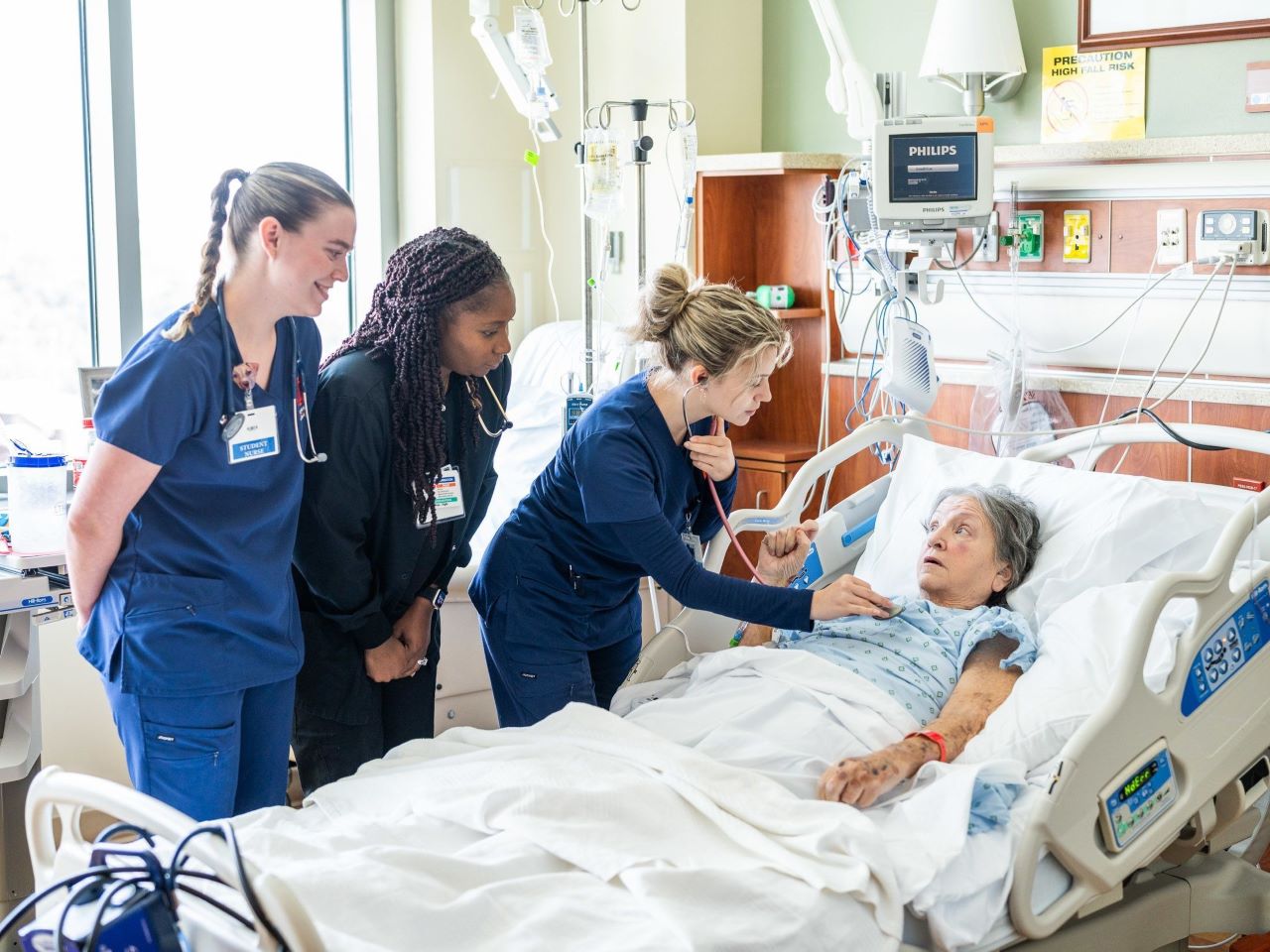 Three nursing students in blue scrubs attend to an elderly woman in a hospital bed