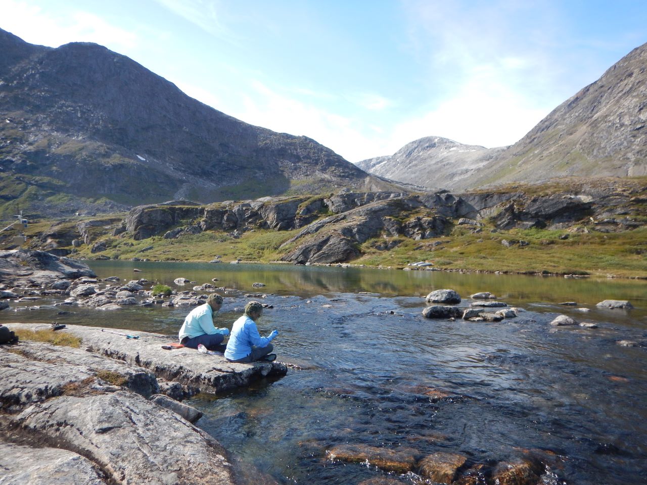 Two scientists sample water from a flowing stream with mountains in the background