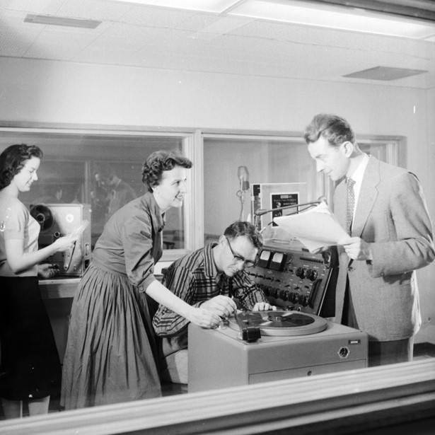 A black and white photo of young men and women gathered around an old-fashioned newsroom